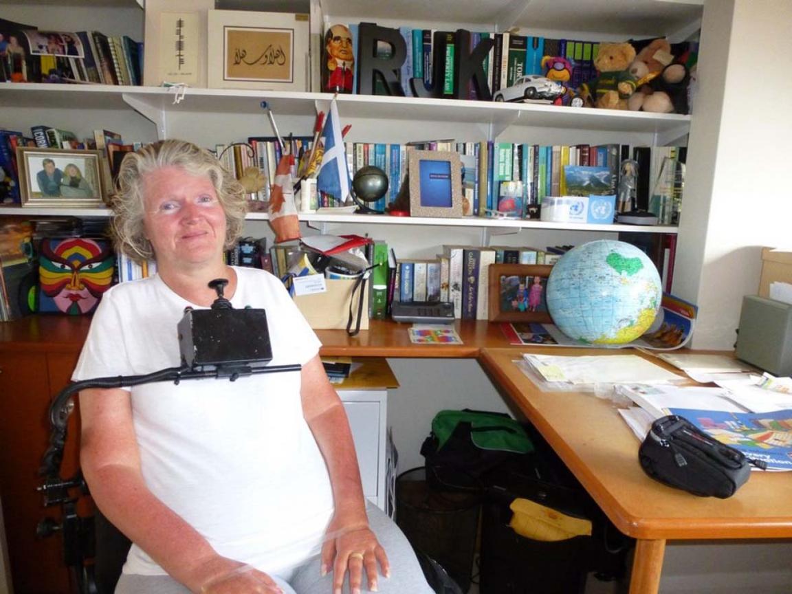 A photograph of a smiling woman seated in a wheelchair in front of a large bookcase and desk. She is wearing a white tshirt. The wheelchair has a control stick which sits just below her chin.