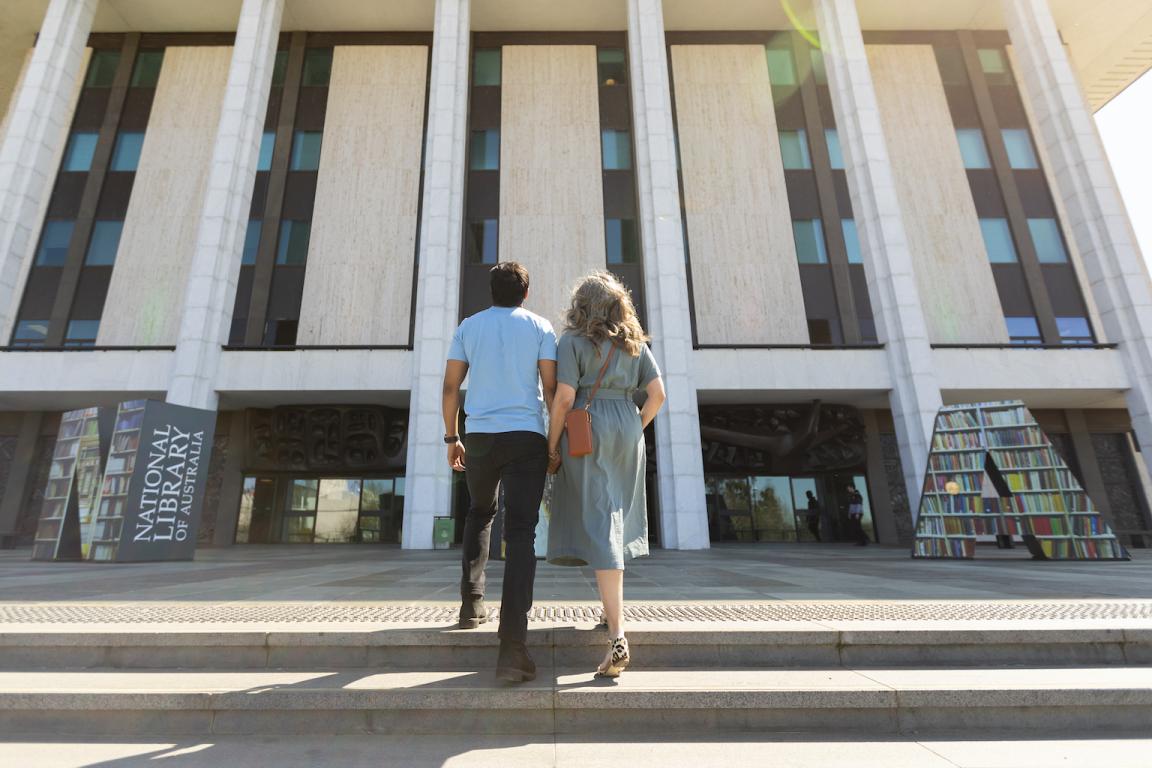 Man and woman in casual wear holding hands and laughing as they walk up the stairs towards the National Library entrance
