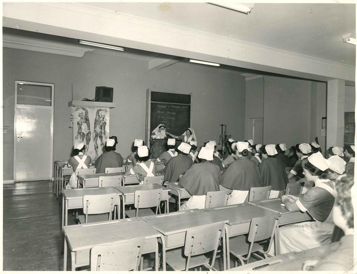 Black and white photo of a classroom full of women in nursing uniforms listening to the teachers at the front of the room who are standing in front of a blackboard and near an anatomical chart