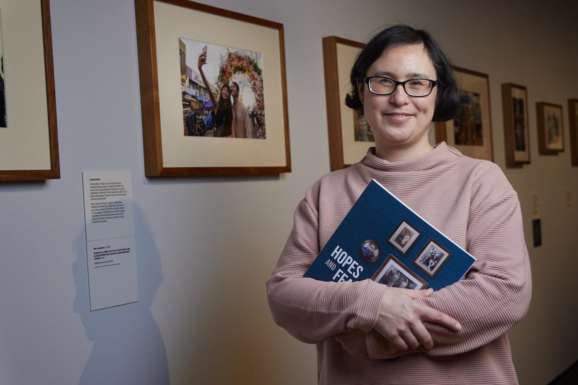 Exhibition curator standing in the Hopes and Fears exhibition gallery holding the exhibition book and smiling