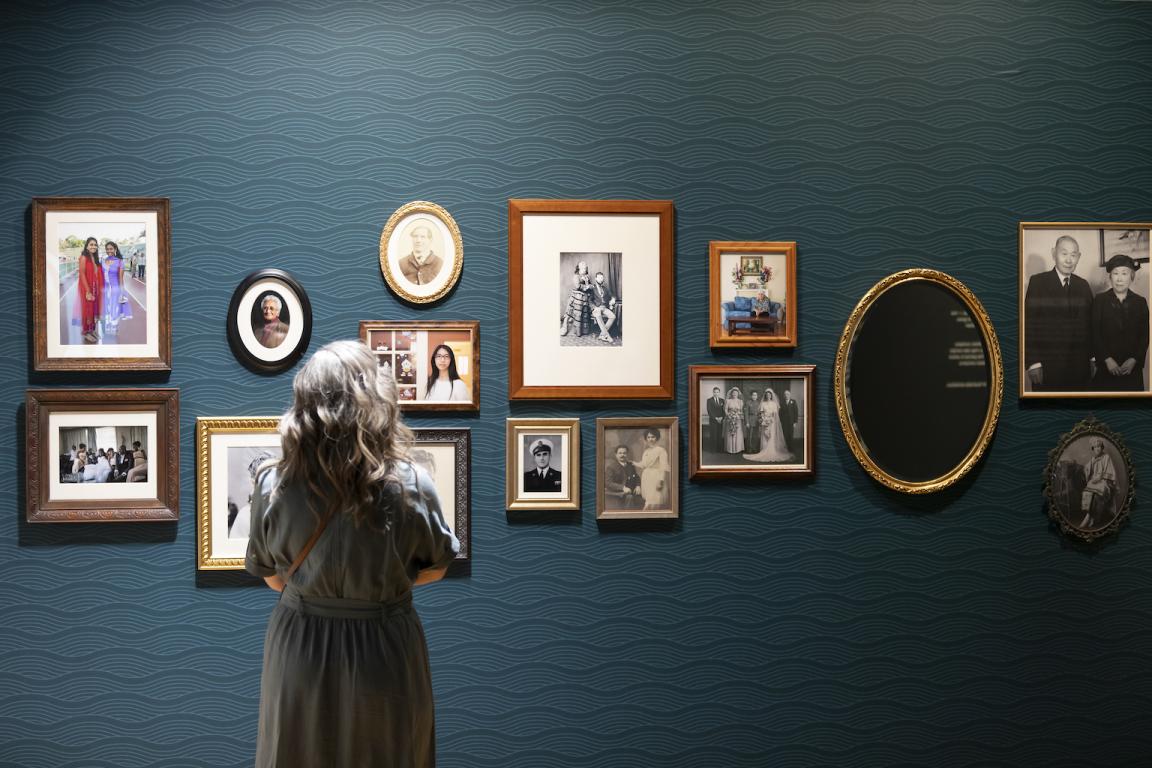 Woman with curly hair standing in front of a wall of framed family photos in the Hopes and Fears exhibition