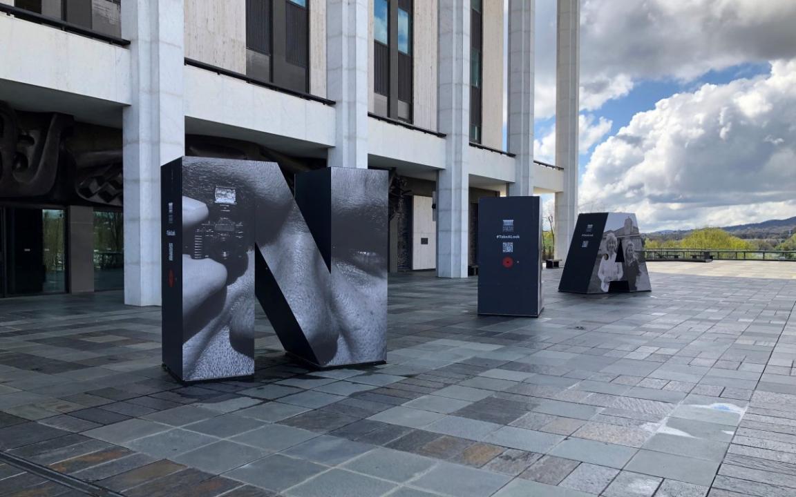 The front podium and colonnade of the National Library of Australia, taken on an angle. On the podium are three huge letters: an 'N', an 'L' and an 'A'. The letters have large photographs on them.