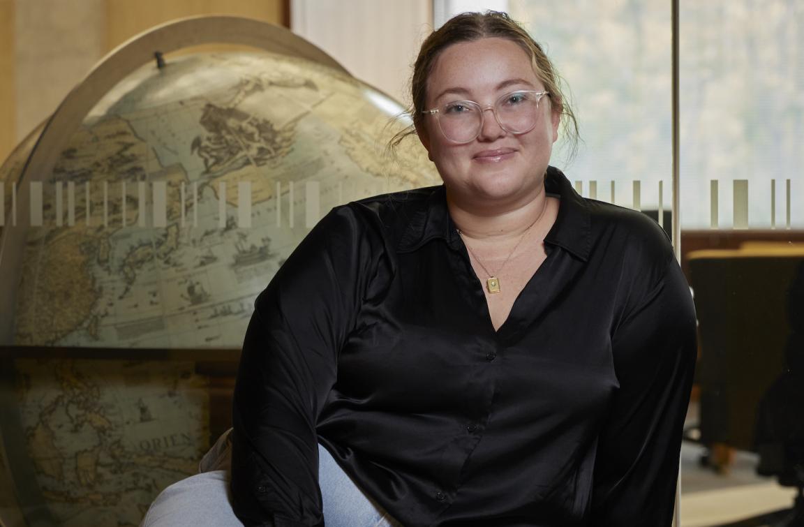 Woman, Jacinta Mackay, sitting in front of a window looking into the Main Reading Room with a large globe on the other side