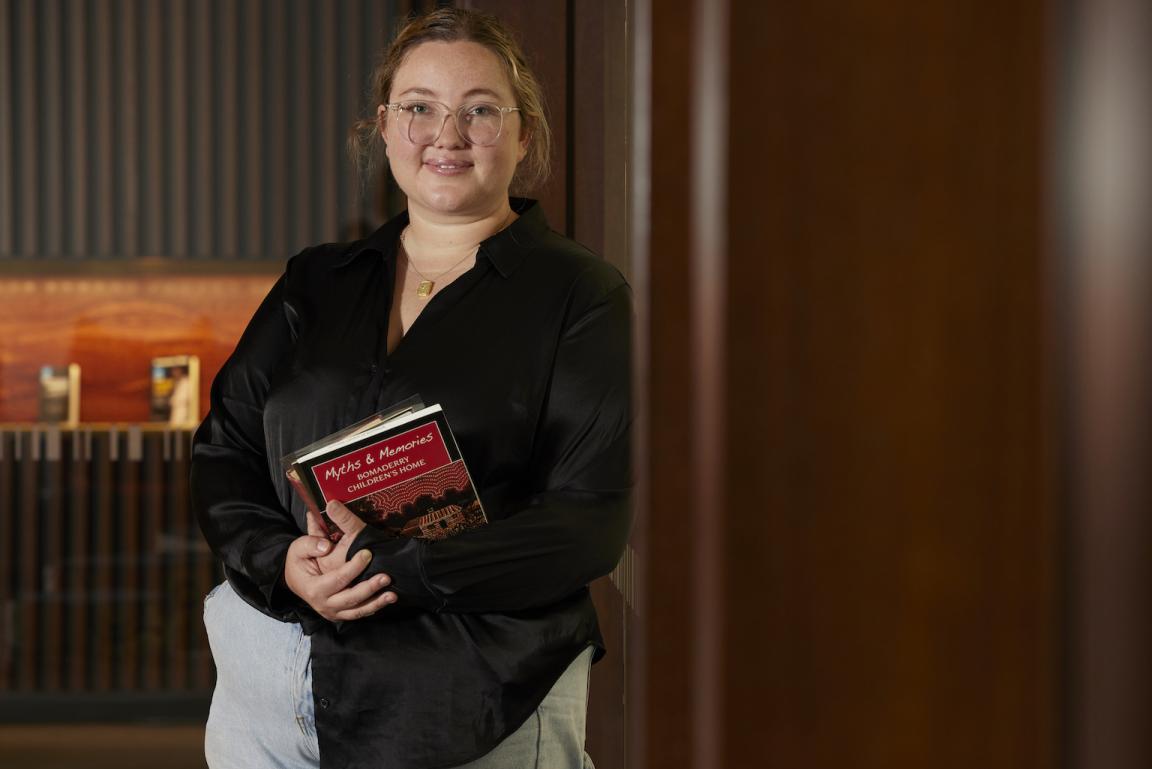 Woman, Jacinta Mackay, leaning against a column in front of the Main Reading Room and holding a book titled 'Myths & Memories: Bomaderry Children's Home'
