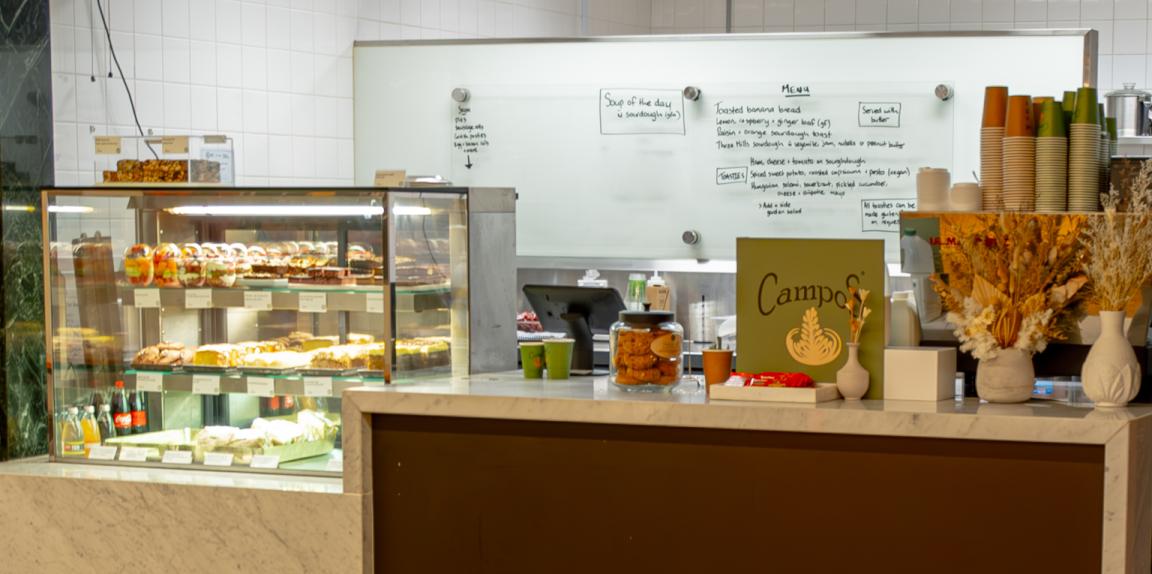 Cafe counter with a display of cakes, slices and fruit salad as well as a coffee machine, stacks of cups and flowers on the counter