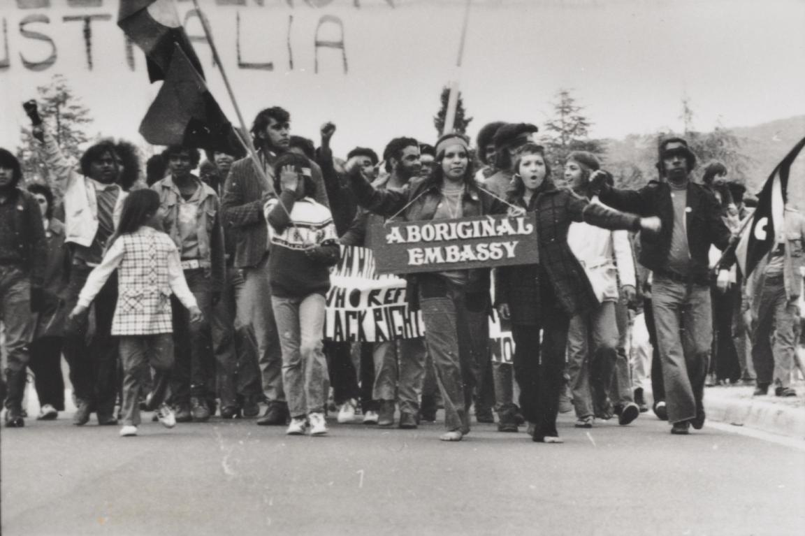 Crowd walking down a road, some with signs and flags. The sign at the front reads 'Aboriginal Embassy'.
