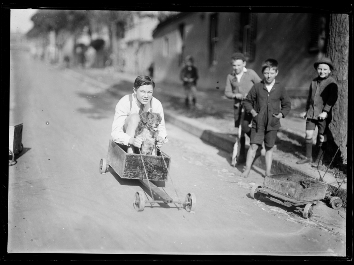 Black and white photo of a boy and dog in a billycart racing down a street