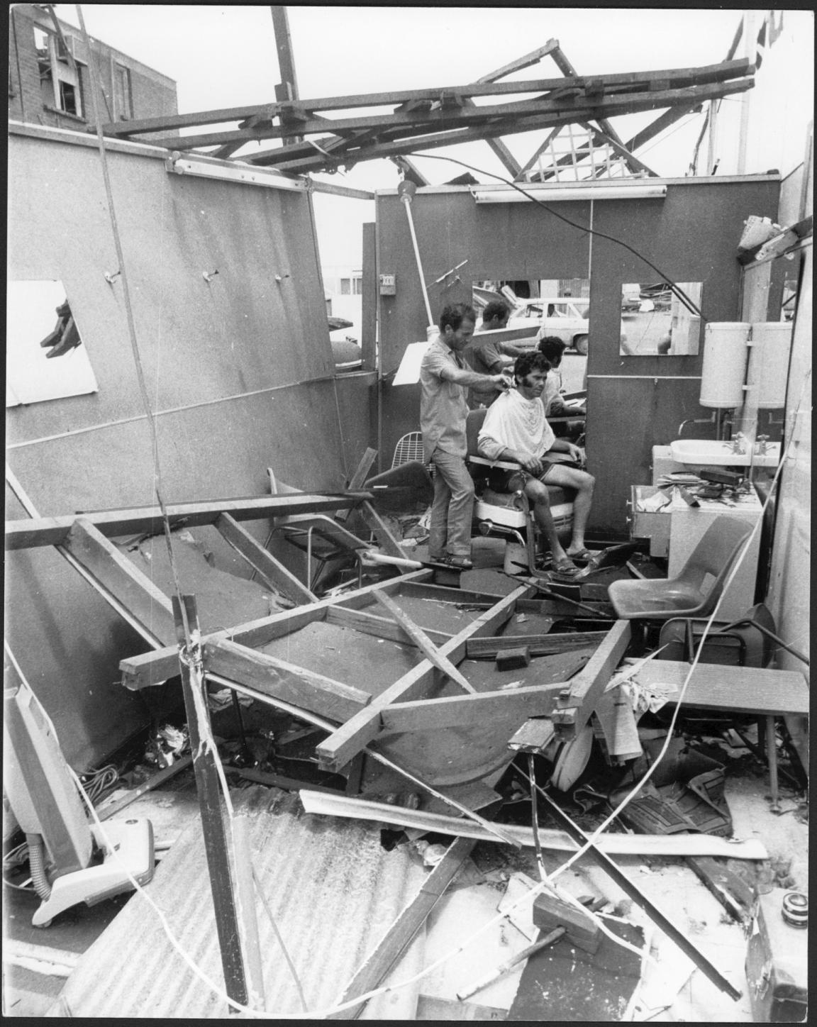 Black and white photo of a barber giving a man a haircut in his shop that was damaged in Cyclone Tracy, with no roof and rubble on the floor 