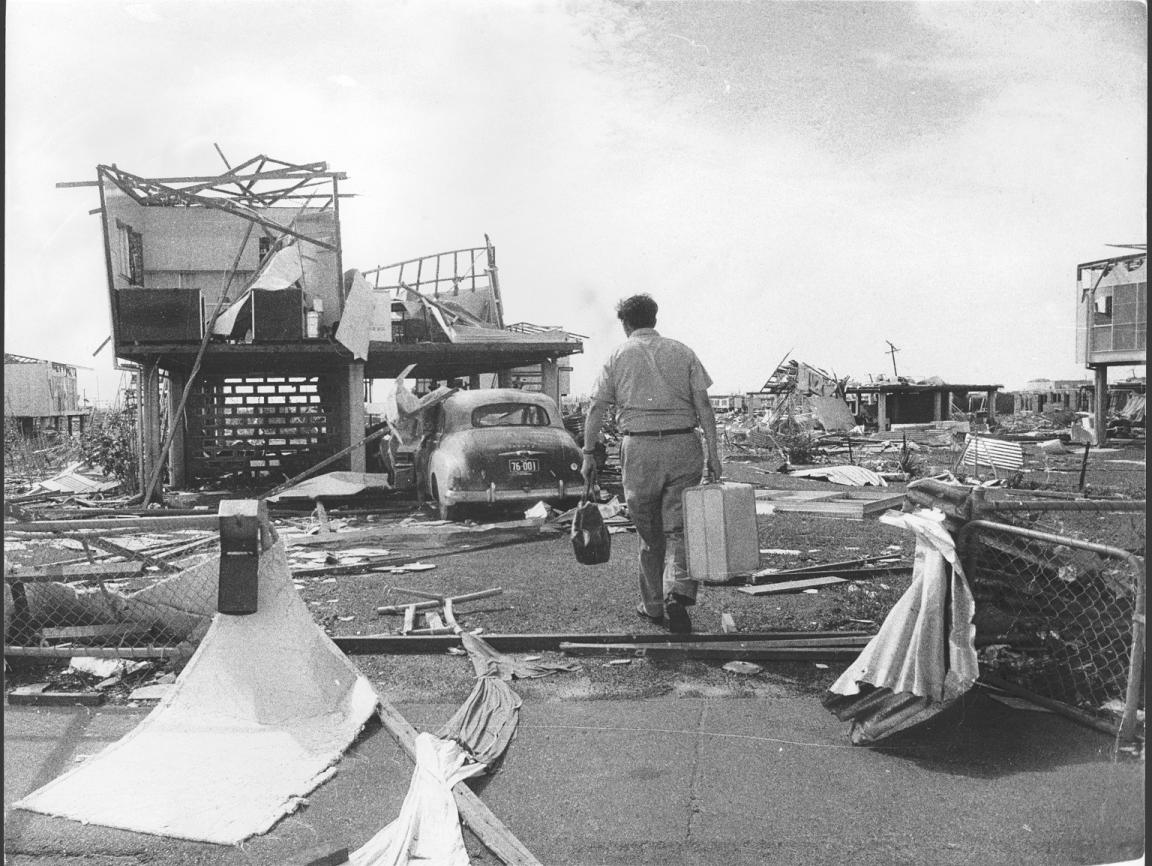 Black and white photo of a man carrying a suitcase walking along a damaged street after Cyclone Tracy