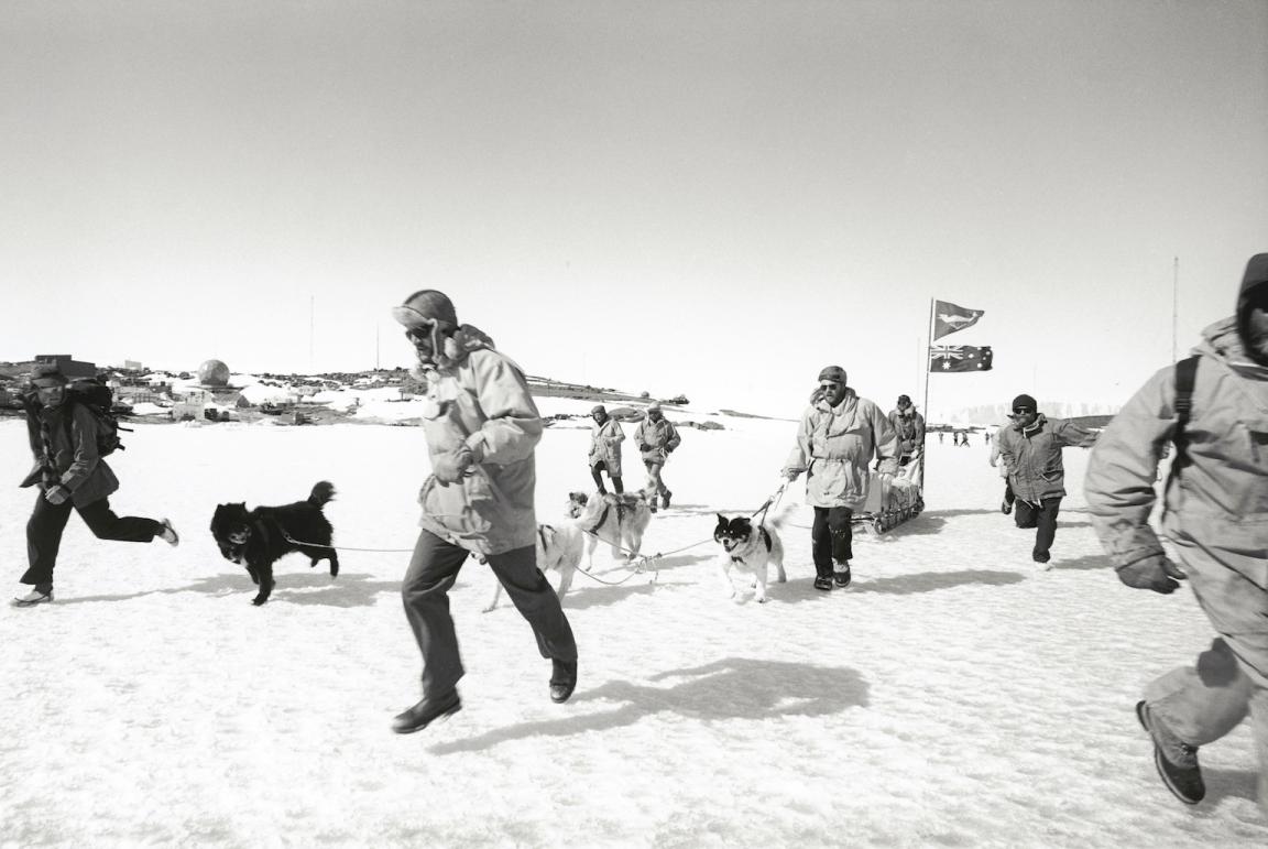 Black and white photo of people in cold-weather gear and huskies running on snowy ground