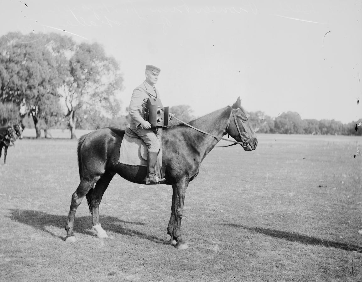 Black and white photo of a man with a large camera around his neck, sitting on a horse