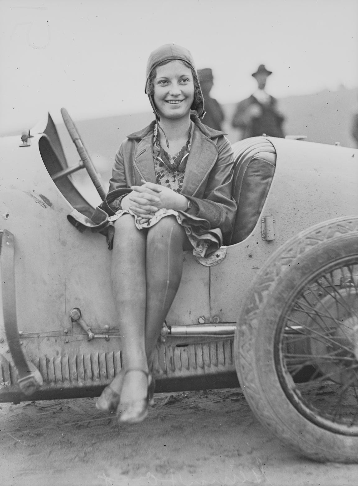 Black and white photo of a woman, Jean Thompson, sitting sideways in an old fashioned race car with a cap and goggles