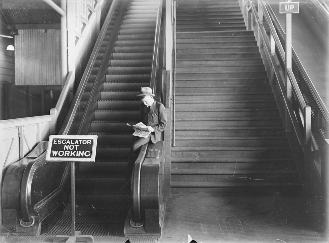 Black and white photo of a man in a suit and hat sitting and reading a newspaper on the handrail of an out-of-order escalator