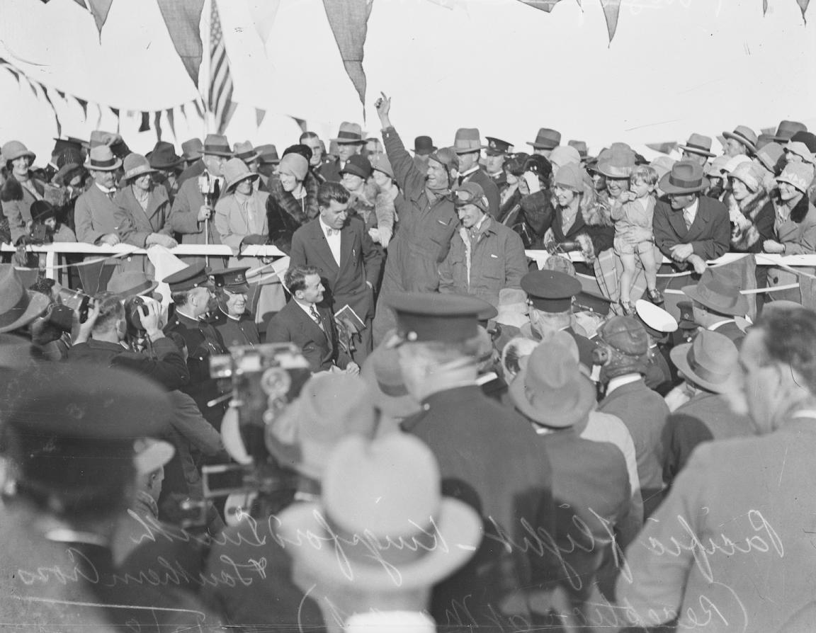 Black and white photo of crowd surrounding a pair of pilots, one of whom has his arm raised in celebration