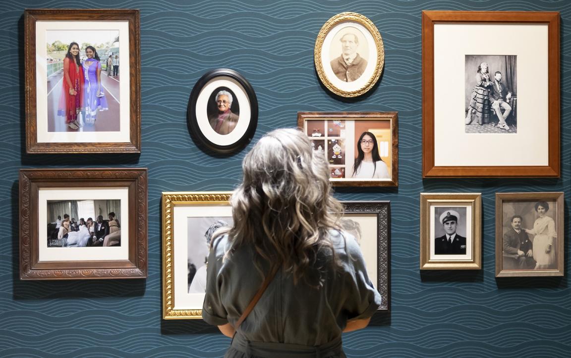 Woman with curly hair standing in front of a wall of framed family photos in the Hopes and Fears exhibition