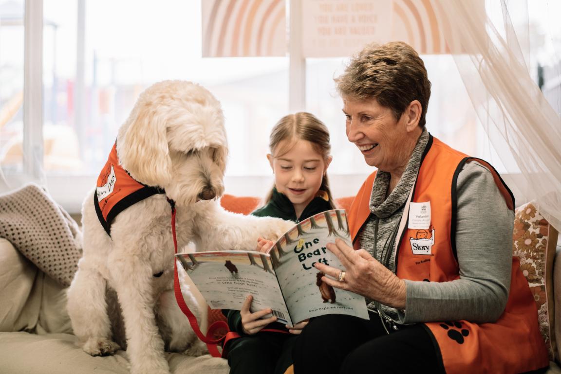 Woman wearing an orange vest reading a book to a young girl with a large white dog sitting next to them with its paw on the book