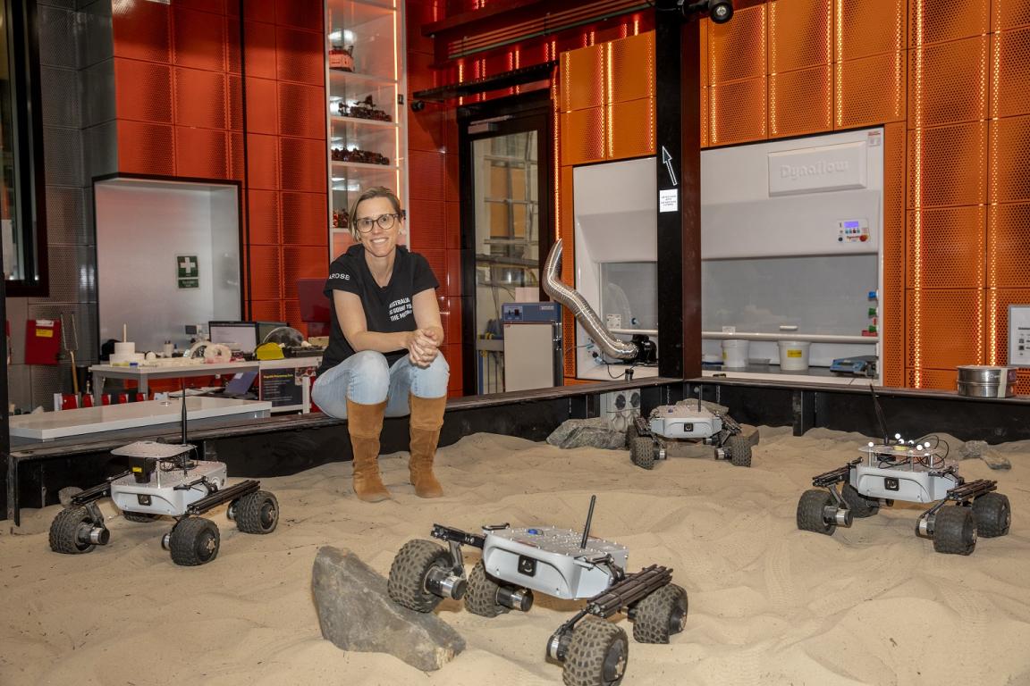Dr Sarah Cannard sitting on the edge of a sand pit with four space rovers in it