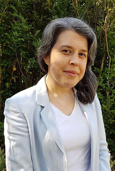 Photo of a woman with greying dark hair, wearing a white shirt and light blue jacket, and standing in front of trees