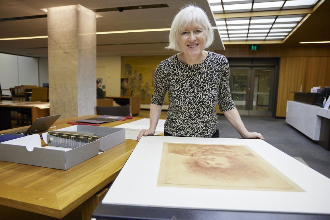 Woman, Dr Christina Thompson, standing in the Special Collections Reading Room, leaning on a table with a large portrait on it and smiling