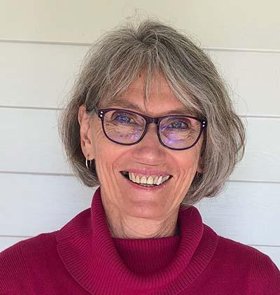 Photo of a smiling woman with grey-brown hair, wearing glasses and a red turtle-neck woollen jumper
