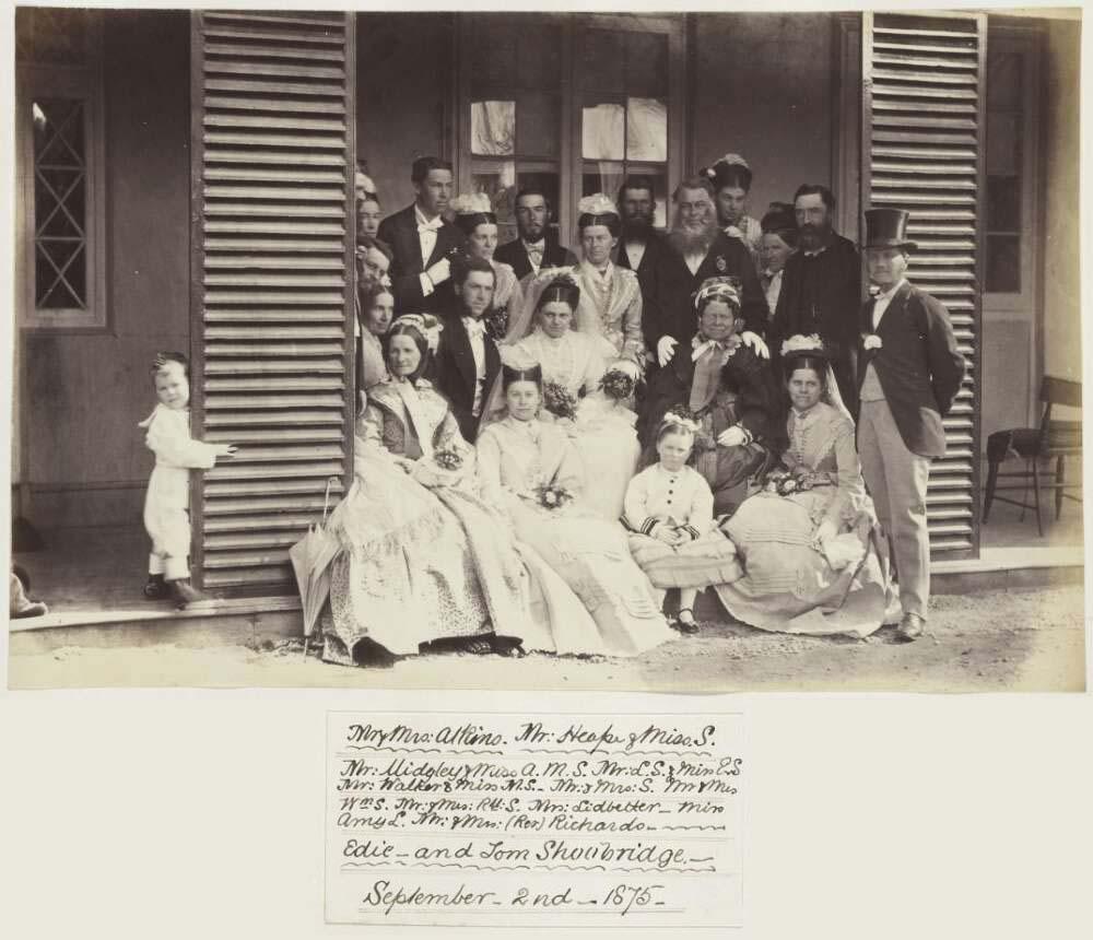 A black-and-white photograph showing a large wedding party posed on the steps of a wooden house. The group includes men, women, and children dressed in formal Victorian attire. The women are in elaborate dresses, some holding small bouquets. The men wear suits, some with top hats, and a small boy peeks around a door frame to the left.