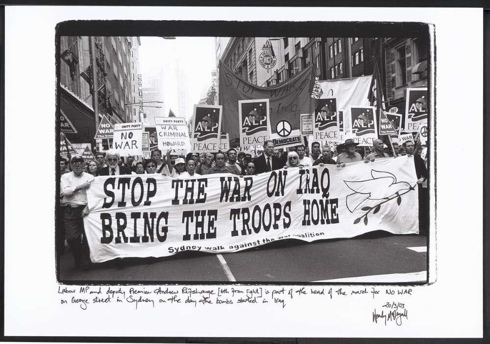 A black-and-white photograph of a large protest march on George Street in Sydney, taken on the day the bombs began in Iraq. The crowd holds a large banner that reads, "STOP THE WAR ON IRAQ BRING THE TROOPS HOME," with a white dove and olive branch illustration on the right. Numerous signs are visible behind the banner, including those from the Australian Labor Party (ALP) and various anti-war groups. Signs include messages such as "NO WAR," "WAR CRIMINAL HOWARD," and "PEACE."