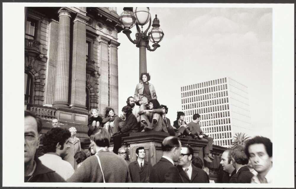 A black and white photo of tens of people gathered on top of and around a large light  on a large stone plinth in front of Victoria's Parliament House.