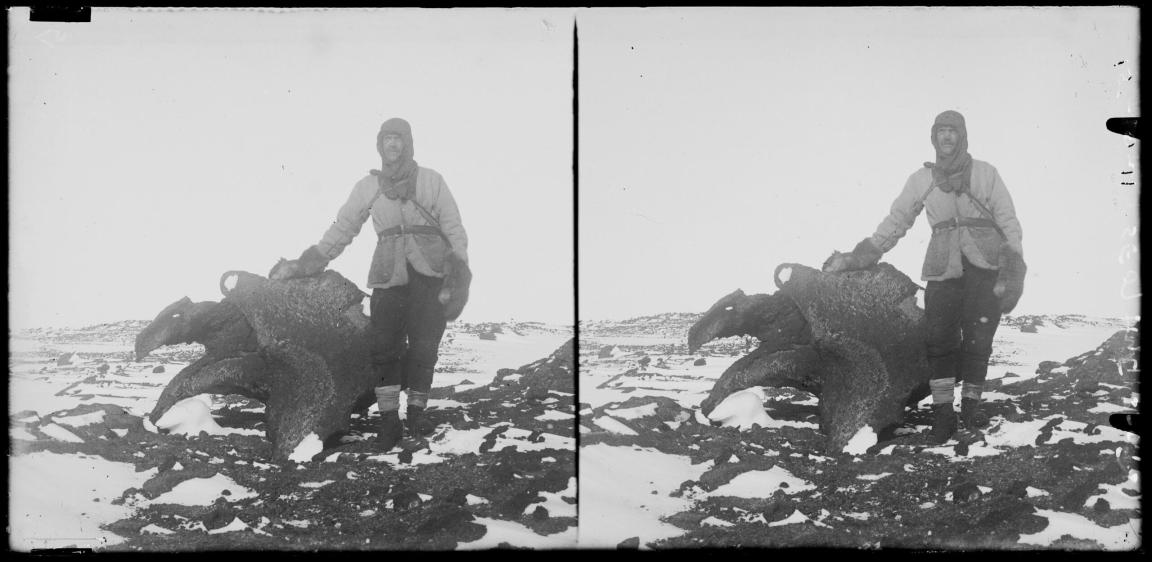 A man in winter attire stands on a plain covered in snow and rocks. He stands beside a large, jagged rock.