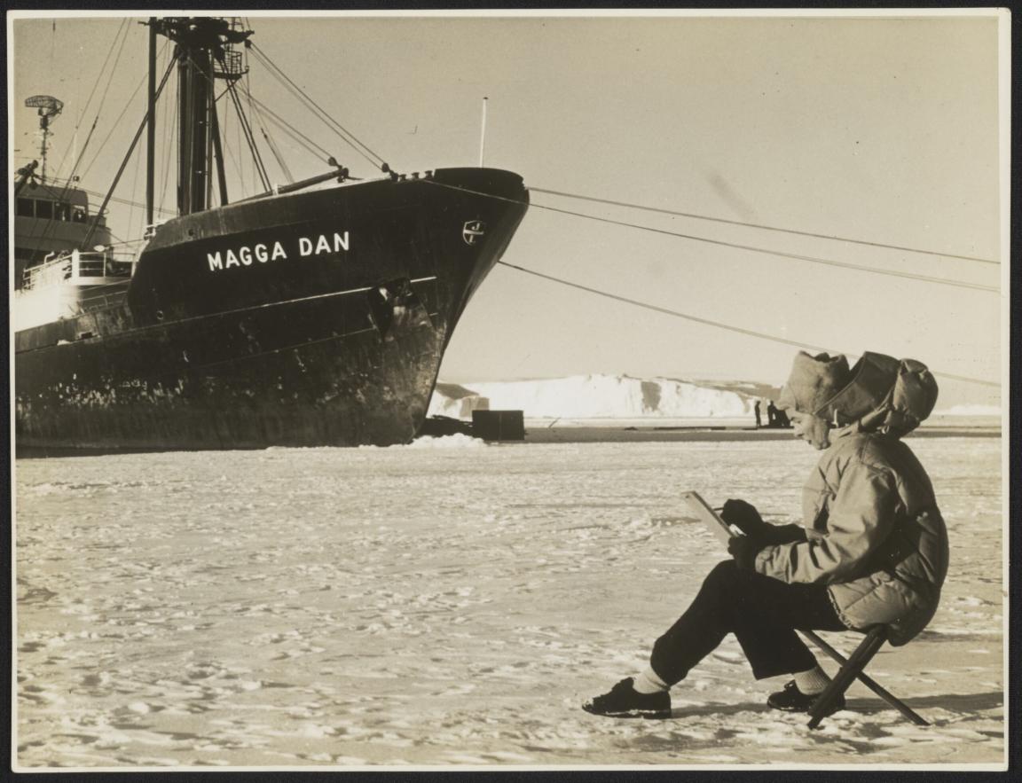A woman sits on a fold-out chair on a snowy plain, sketching in a book. A large icebreaker ship is moored in the background.