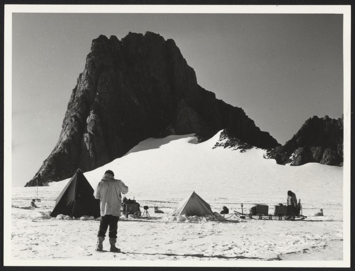A man standing on a snowy plain, in front of tents, sledges with scientific equipment, scientists and sled dogs. A large rocky outcrop looms in the background.