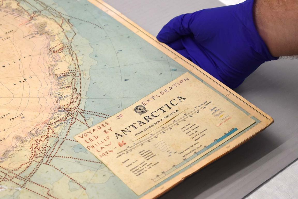 A hand wearing a blue glove holds the bottom right corner of a map of Antarctica including the handwritten words 'Voyages of Exploration Led By Phillip Law 1954-66'