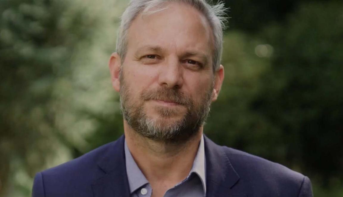A man with grey hair and brown and grey beard and moustache standing in front of blurred out trees
