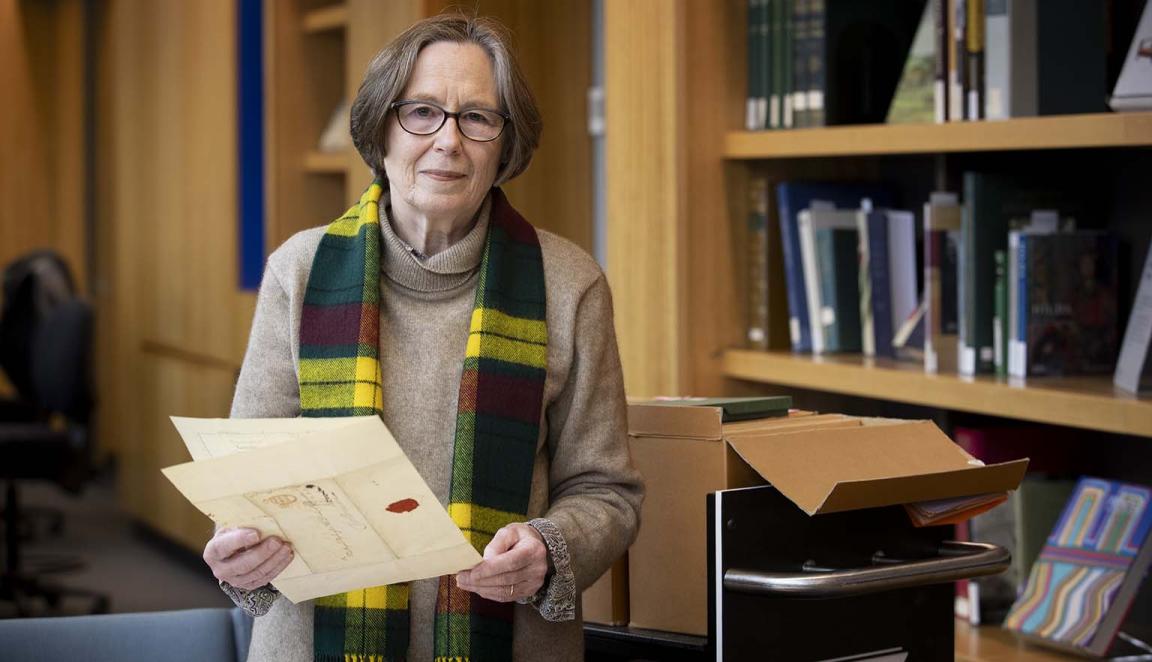 A woman with short hair and glasses smiles and looks at the camera while she holds a folded document in her hands. In the background are bookshelves with books and an open box on a trolley.