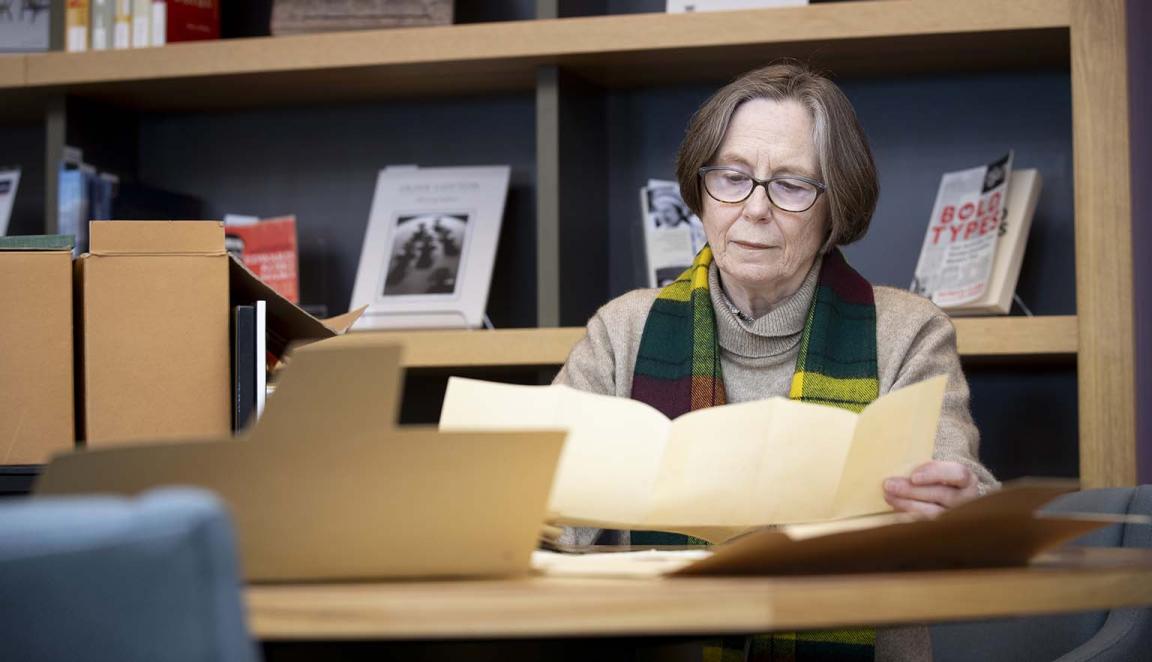 A woman with short hair and glasses sits at a desk looking at a yellowed piece of paper she holds in her hands