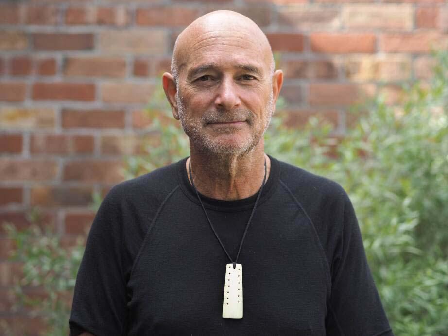 A man stands in front of a brick wall and a bush.