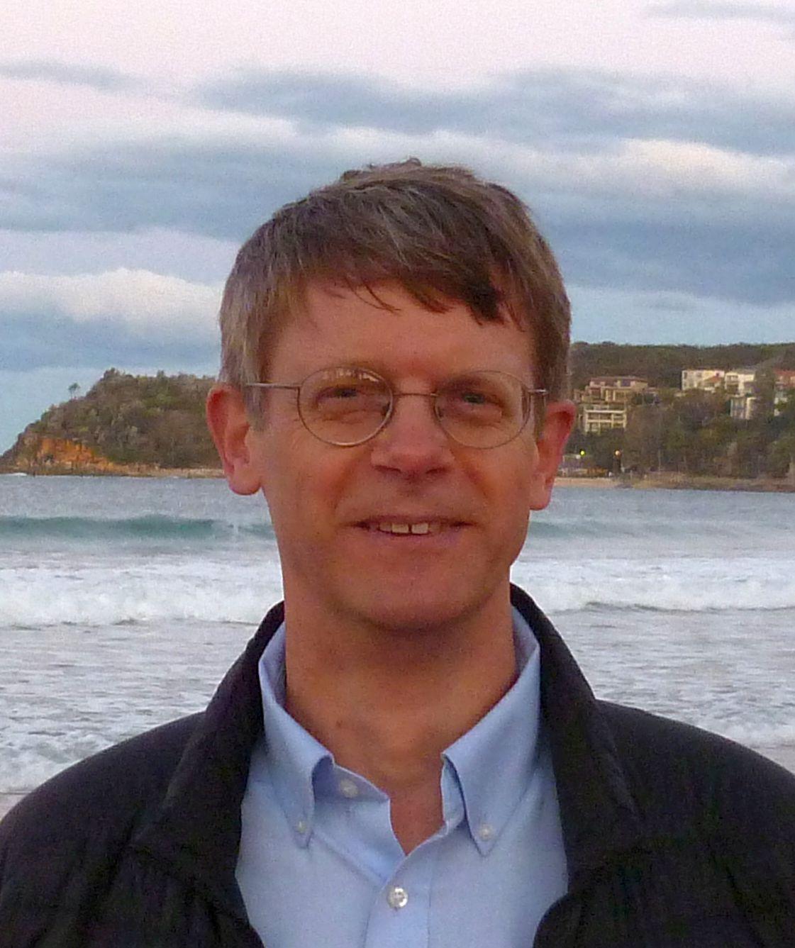 Chet Van Duzer in a blue shirt and jacket standing in front of the ocean