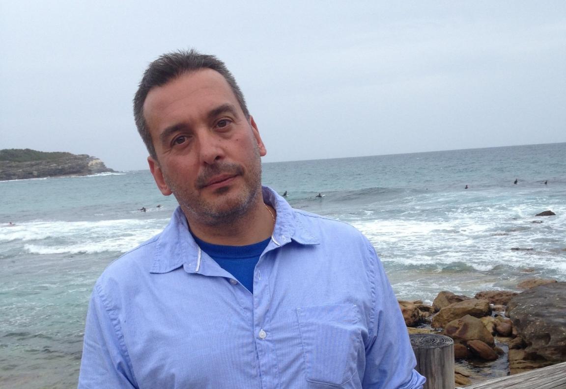 Christos Tsiolkas wearing a blue shirt standing in front of the ocean by a rock pool