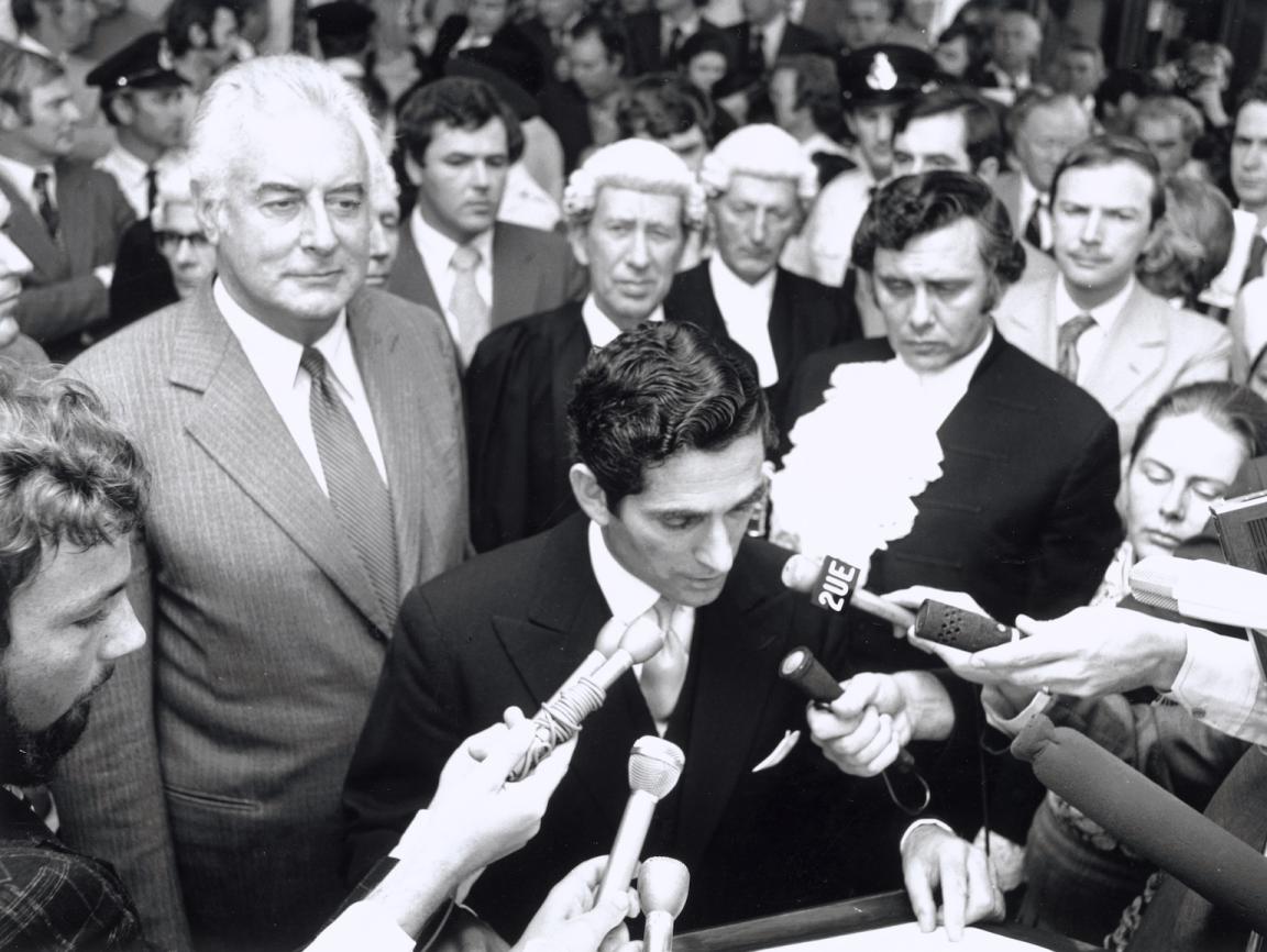 Black and white photo of Mr David Smith speaking at a podium with several microphones held up in front of him with Gough Whitlam standing behind him in the crowd