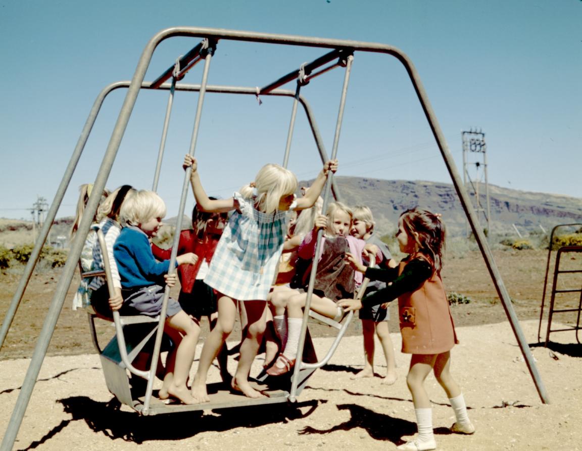 Photo of several young children in 1970s-era clothing playing on a swing set on a sunny cloudless day