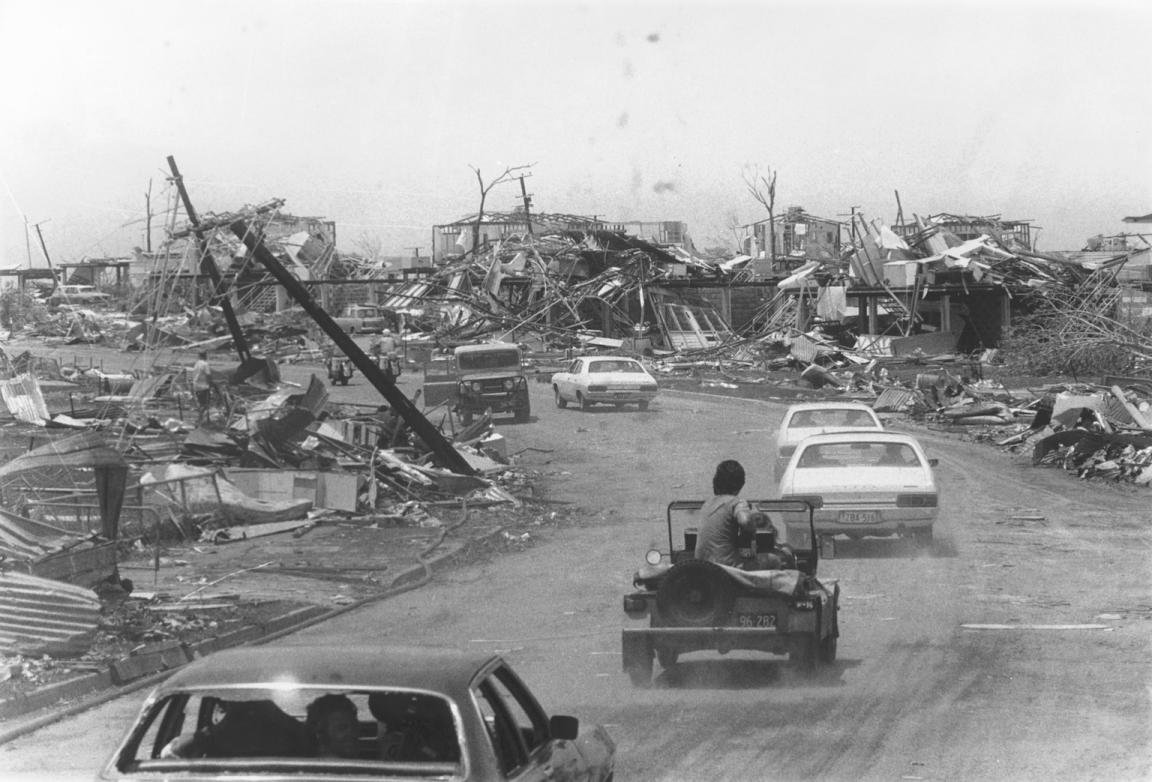 Black and white photo of cars driving a dirty road lined with wreckage from Cyclone Tracy