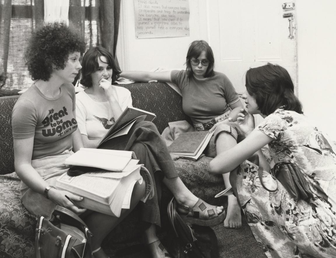 Black and white photo of three women sitting on a couch and a woman crouching in front of them