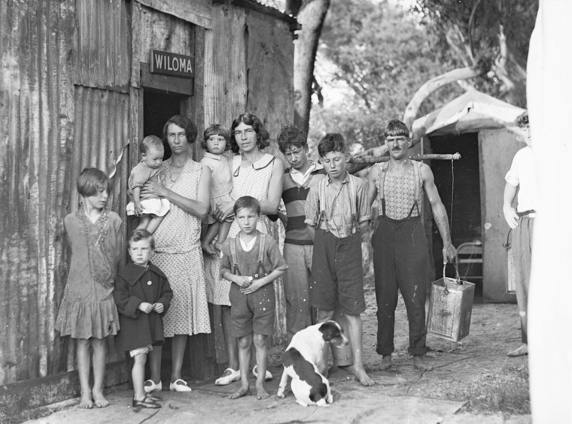 Black and white photo of a large family with many children and a dog standing outside a tin shack