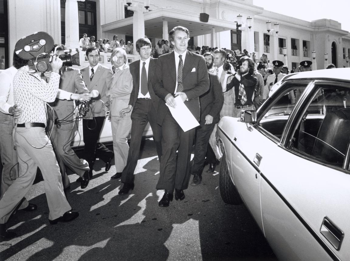 Black and white photo of Malcolm Fraser walking in front of Parliament House followed by a crowd including journalists and photographers
