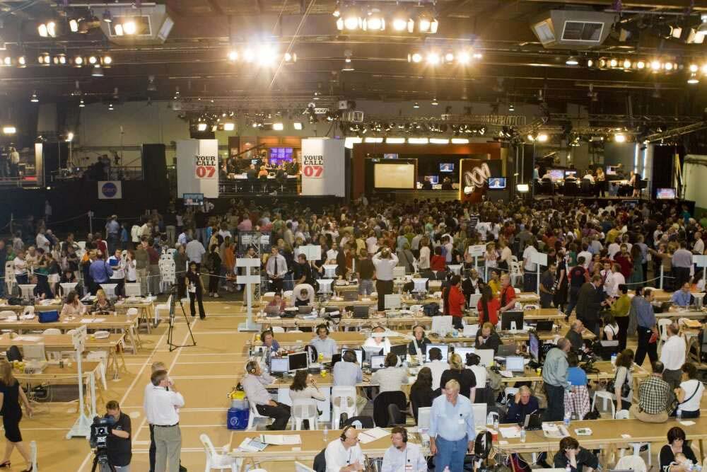 People standing, sitting at desks, and filming with news cameras in a brightly lit room