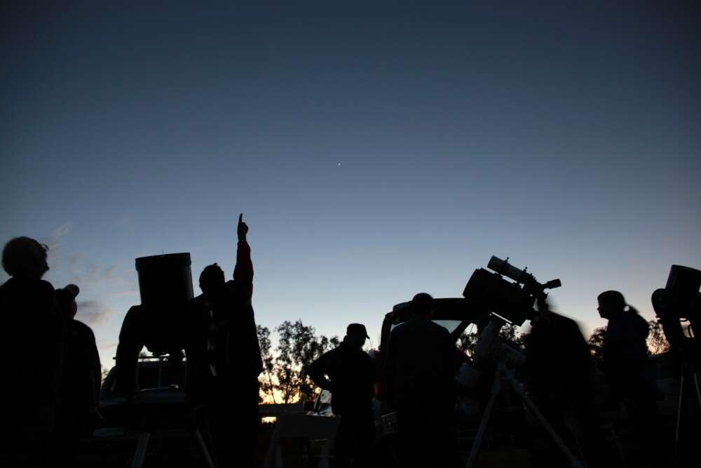 A group of people at dusk standing beside small telescopes and pointing at the sky.