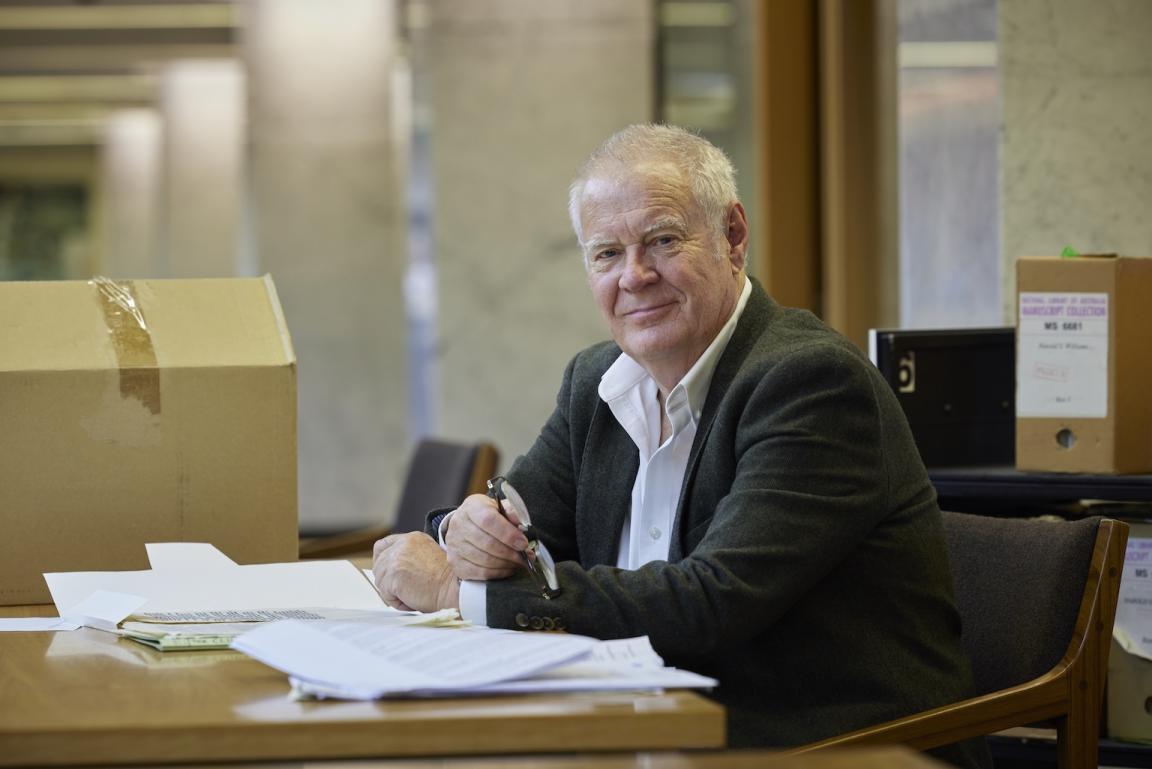 Man, Professor Robin Gerster, smiling and sitting at a table in the Special Collections Reading Room with manuscipt material in front of him