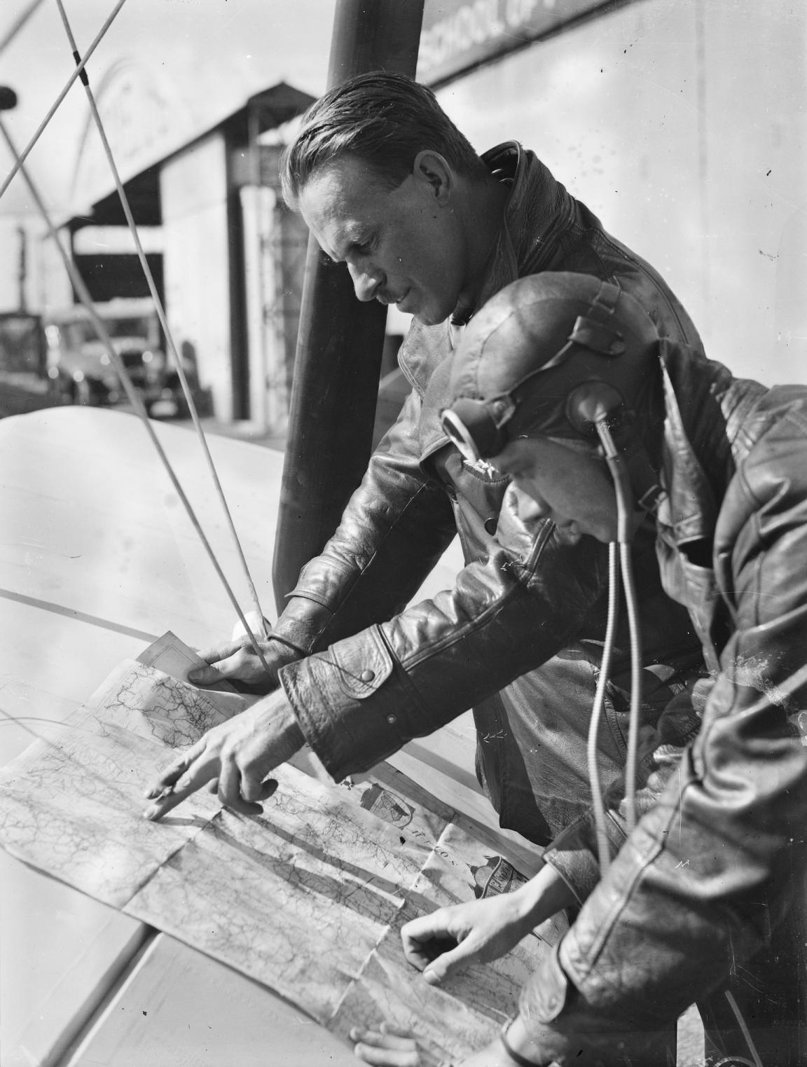 Black and white photo of two aviators looking and pointing at a map laid over the wing of a small plane