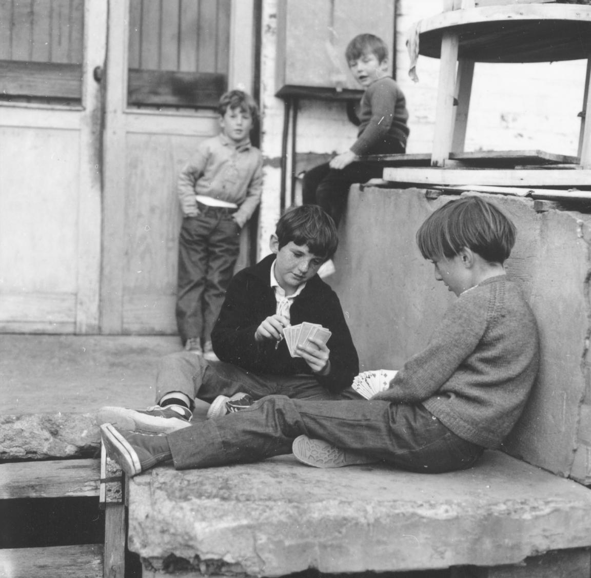 A black and white photograph of two boys playing cards while another two boys are watching