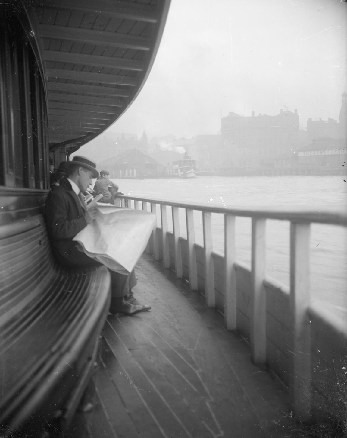 Black and white photo of a man sitting on the deck of a ferry reading a newspaper