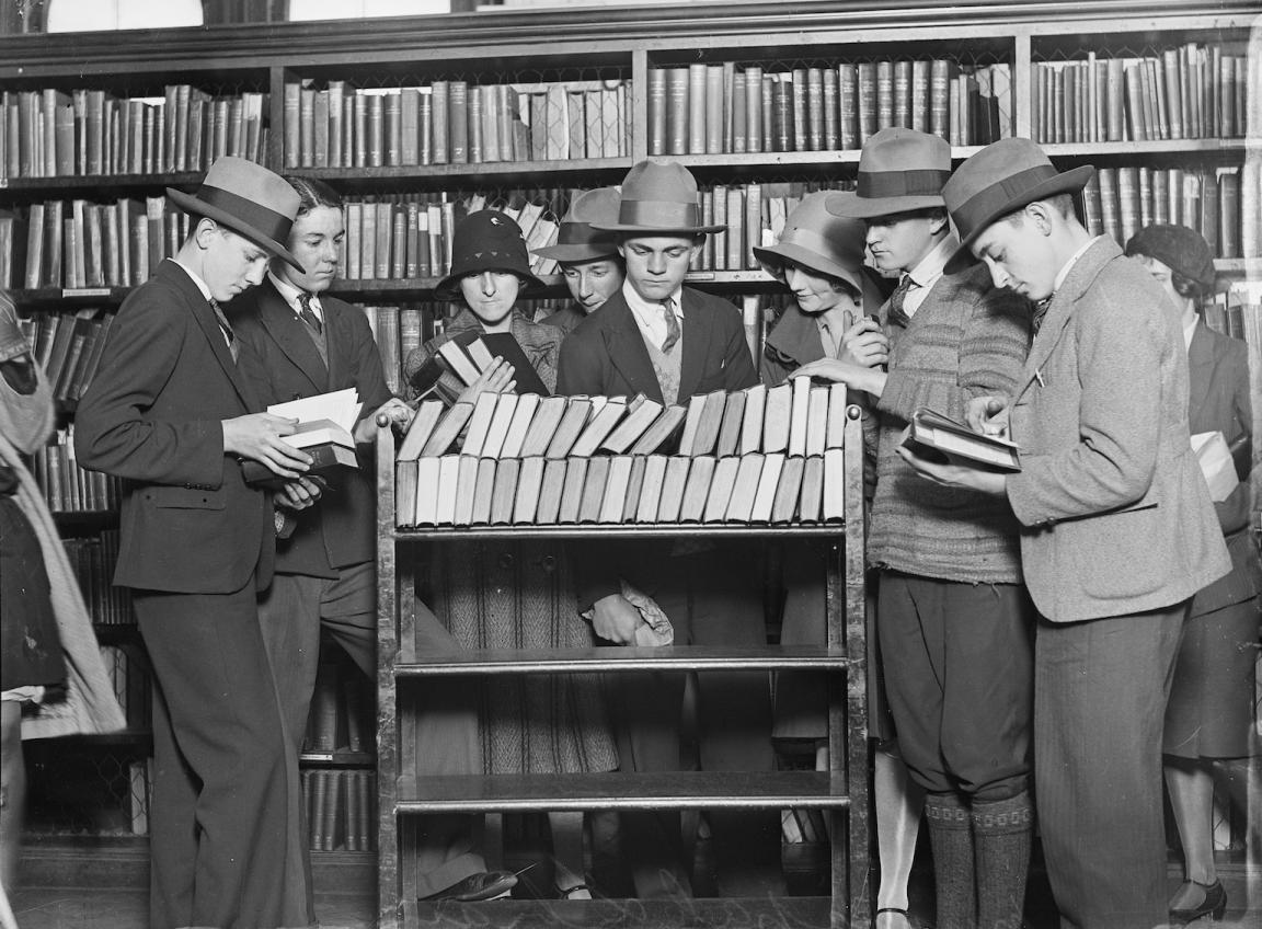 Black and white photo fo a group of men and women gathered around a trolley full of books in a library